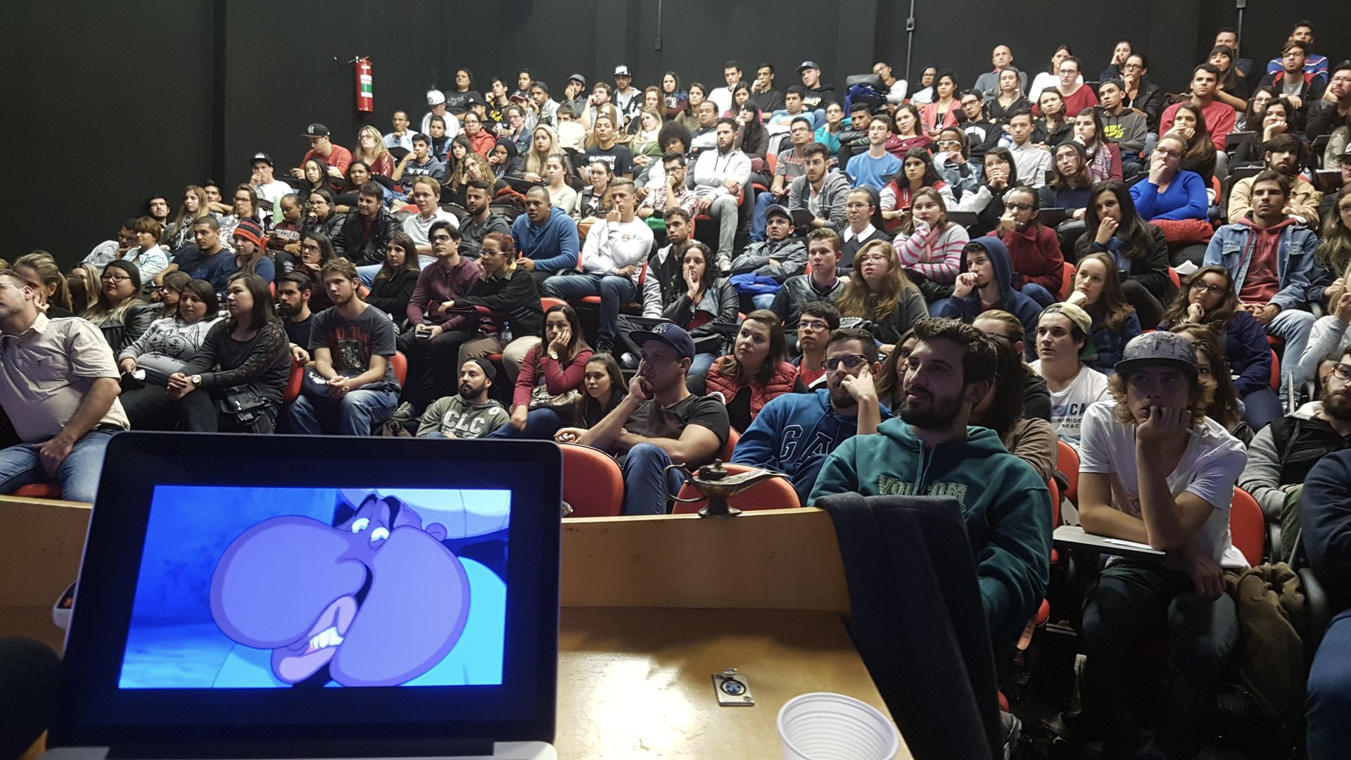 Vista do palco para arquibancada lotada de estudantes, monitor em primeiro plano com o gênio do Aladdin