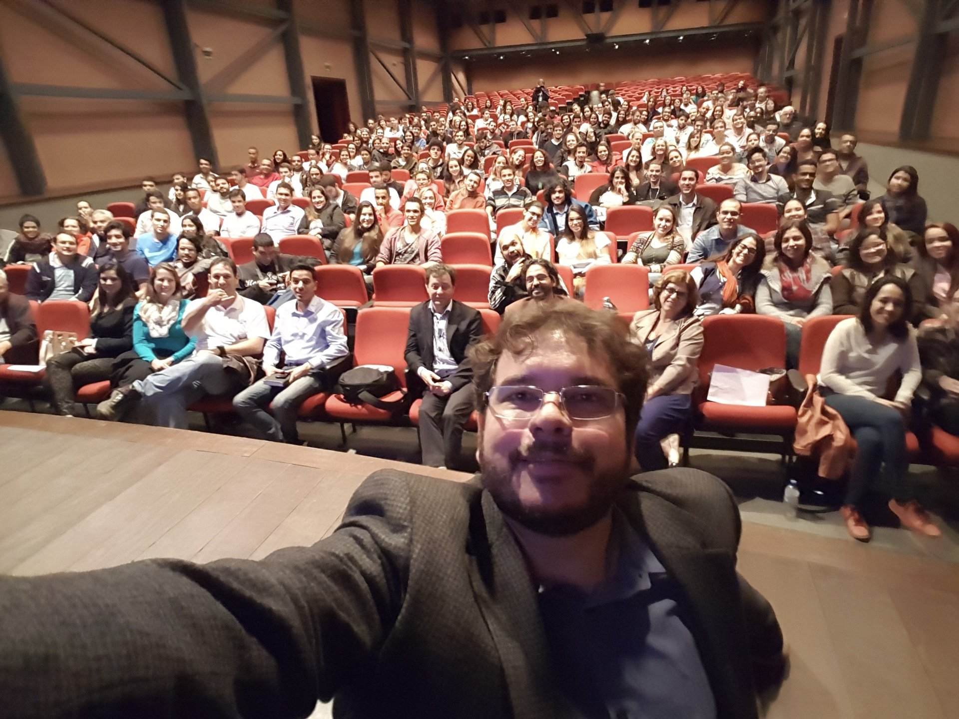 Selfie de homem de blazer em palco de madeira com auditório de poltronas vermelhas lotado atrás