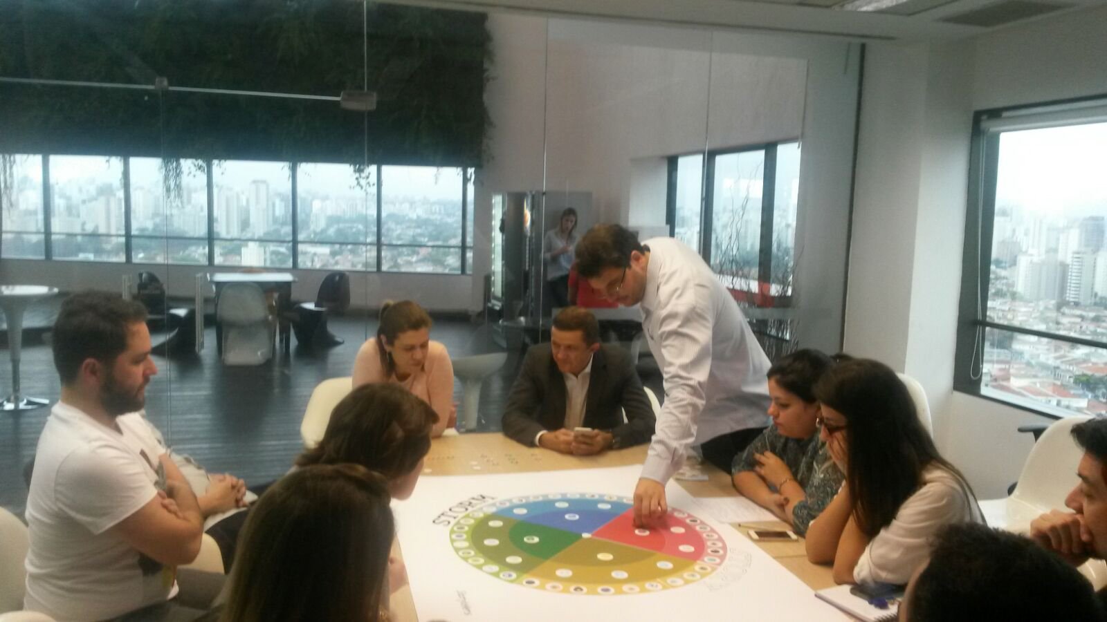Homem apresenta em sala envidraçada com o tabuleiro Storytellers na mesa e o skyline de São Paulo ao fundo